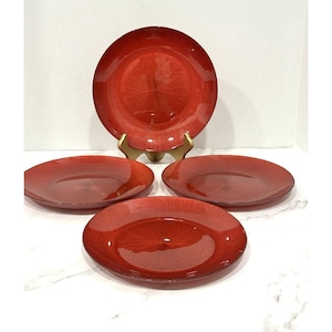 May include: Set of four red glass plates with a textured, radial design. One plate is displayed on a gold-colored stand, while the other three are arranged in front. The plates have a glossy finish and a slightly raised edge.
