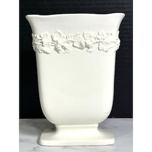 May include: A white ceramic vase with a flared rim and a square base. The upper portion of the vase features a raised, embossed relief pattern depicting grapevines, leaves, and clusters of grapes encircling the entire circumference of the vessel.