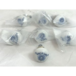 May include: A collection of white ceramic cabinet knobs with blue floral designs. Each knob is individually wrapped in clear plastic. The knobs have a rounded shape and a silver-colored metal base.