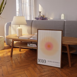 May include: A framed print with a blurred, peach and orange circle in the center. The print has the text "1010 DIVINE GUIDANCE" at the bottom. The print is sitting on a wooden table with a lamp and books.