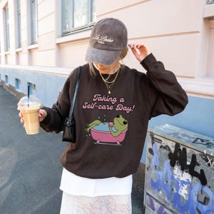Könnte beinhalten: Brauner Pullover mit einem grünen Frosch in einer Badewanne und dem Text "Taking a Self-care Day!"