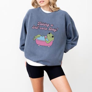 Könnte beinhalten: Ein blaues Sweatshirt mit dem Text "Taking a Self-care Day!" und einem Cartoon-Frosch in einer Badewanne. Der Frosch ist grün mit rosa Augen und hält eine rosa Tasse. Die Badewanne ist rosa mit weißen Blasen.