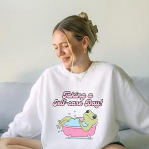 Könnte beinhalten: Weißer Pullover mit einem Aufdruck einer grünen Frosch in einer rosa Badewanne mit dem Text "Taking a Self-care Day!" in rosa Schrift.