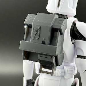 May include: A white action figure of a Star Wars clone trooper wearing a black backpack with a raised rectangular design. The backpack has a raised symbol on the front.