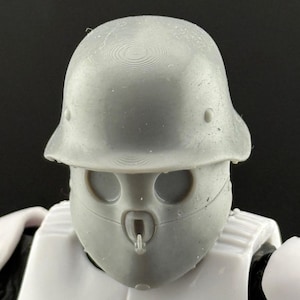 May include: Close-up of a gray helmet and face mask, part of a white and black action figure. The helmet has a rounded top with a slight brim. The face mask has two eye openings and a central breathing apparatus.