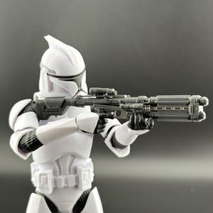以下が含まれることがあります： 黒いブラスターライフルを持った白いクローン・トルーパーのアクションフィギュア。フィギュアは、黒いアクセントとヘルメットが付いた白いアーマーを着用しています。背景は無地のグレーです。
