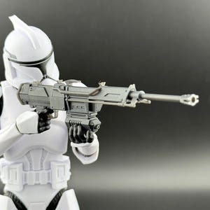 May include: A white action figure of a clone trooper holding a black blaster rifle. The figure is wearing white armor with a white helmet.