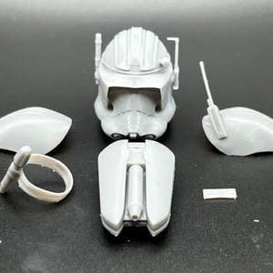 May include: A disassembled, light gray model kit of a helmet and related components. The kit includes the helmet's main structure, side pieces, and other small parts, all arranged on a dark surface.