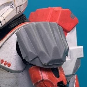 May include: A close-up of a Star Wars action figure wearing a white and red armor with a gray shoulder pad. The figure is holding a blaster rifle.