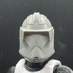 May include: A white and gray plastic clone trooper helmet with a visor. The helmet has a textured surface and a raised design on the top.