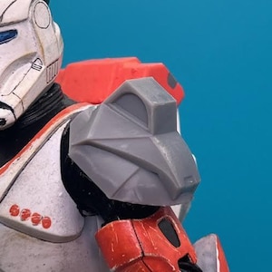 May include: A close-up of a gray and red action figure with a helmet. The figure is wearing a white and red suit with a gray shoulder pad.