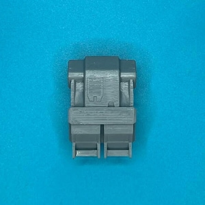 May include: A gray plastic connector with two rectangular openings. The connector has a textured surface and a small rectangular indentation on the top.