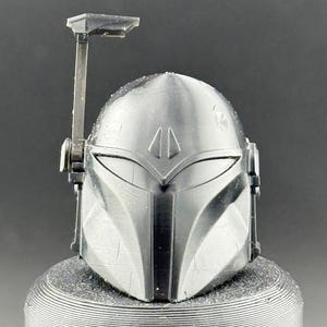 K&ouml;nnte beinhalten: Ein silberner Mandalorianer-Helm mit einem T-f&ouml;rmigen Visier und einem Entfernungsmesser. Der Helm mit seinem kantigen Design steht auf einem strukturierten, zylindrischen Sockel. Das Design ist von der Star Wars-Franchise inspiriert.