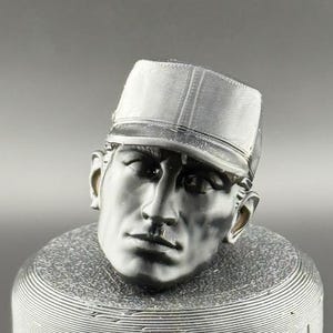 Peut inclure: Une sculpture argentée représentant une tête d'homme portant une casquette. La sculpture est montée sur une base cylindrique avec des rainures horizontales. Le visage de l'homme présente des traits détaillés, dont un nez, des lèvres et des yeux bien définis. La casquette a une visière.