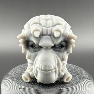 May include: A 3D printed gray alien head with a detailed face and textured skin. The head is on a gray cylindrical base.