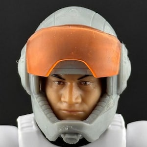 以下が含まれることがあります： グレーのヘルメットとオレンジ色のバイザーが付いたフィギュアの頭と肩のクローズアップ。フィギュアはニュートラルな表情で、顔の特徴が見えます。フィギュアは白い制服を着ています。