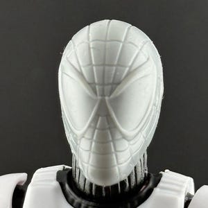 May include: A close-up of a white action figure head with a Spider-Man mask design. The mask has a webbed pattern and large eye openings. The figure's body is white with black accents around the neck.