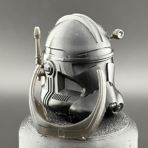 May include: A black plastic Star Wars clone trooper helmet with a visor and a small antenna on top. The helmet is sitting on a gray surface.