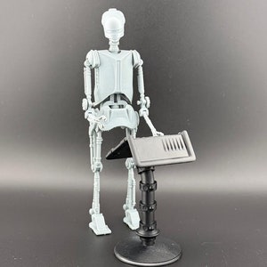 May include: A gray plastic action figure of a robot standing next to a black plastic stand with a flat surface and a small control panel. The robot has a head, torso, arms, and legs.