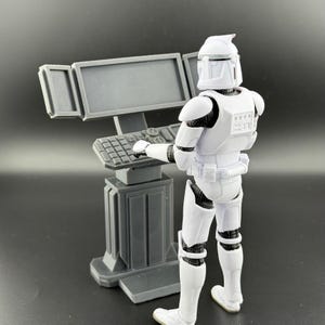 May include: A white action figure of a stormtrooper standing in front of a grey computer console with a keyboard and a monitor. The stormtrooper is holding his right hand on the keyboard.