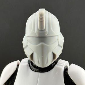 May include: A close-up of a white and grey helmet, part of a costume. The helmet has a distinctive design with a visor and a raised central ridge. The figure is wearing a white chest piece and a black undersuit. The background is a solid grey.