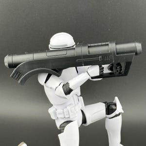 May include: A white action figure of a Star Wars Stormtrooper holding a black blaster rifle. The rifle has a scope and a handle.