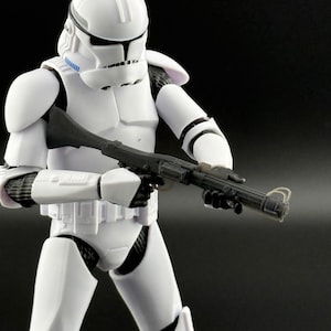 May include: A white action figure of a Star Wars clone trooper holding a black blaster rifle. The figure has a white helmet with a blue visor and a white and black chest plate.