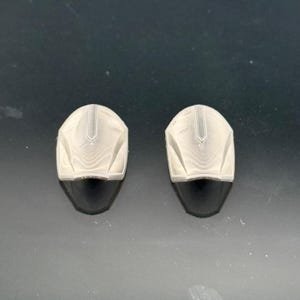 Pu&ograve; includere: Due piccoli oggetti a forma di casco, di colore chiaro, con una visiera scura e curva. I caschi hanno una superficie strutturata con una cresta centrale e sono posti su uno sfondo scuro.
