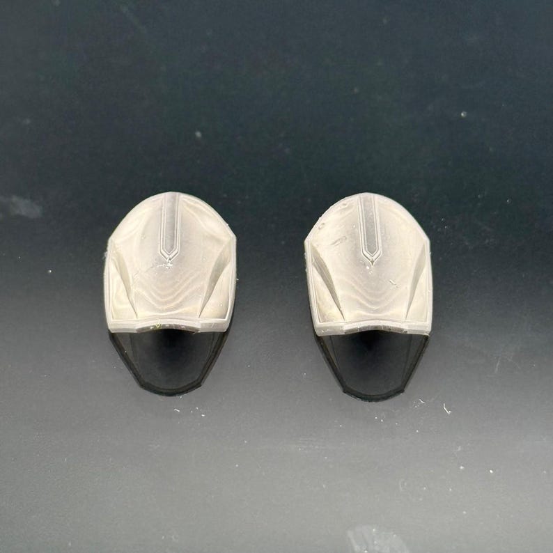 Pu&ograve; includere: Due piccoli oggetti a forma di casco, di colore chiaro, con una visiera scura e curva. I caschi hanno una superficie strutturata con una cresta centrale e sono posti su uno sfondo scuro.