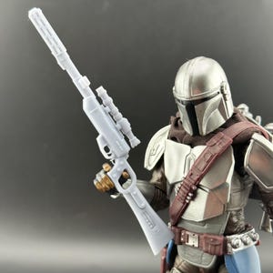 Puede incluir: Una figura de acci&oacute;n mandaloriana gris claro que sostiene un rifle gris claro con mira telesc&oacute;pica. La figura lleva un casco plateado, una bufanda marr&oacute;n y una placa pectoral marr&oacute;n y plateada. La figura tiene un cintur&oacute;n marr&oacute;n y un atuendo azul y marr&oacute;n.