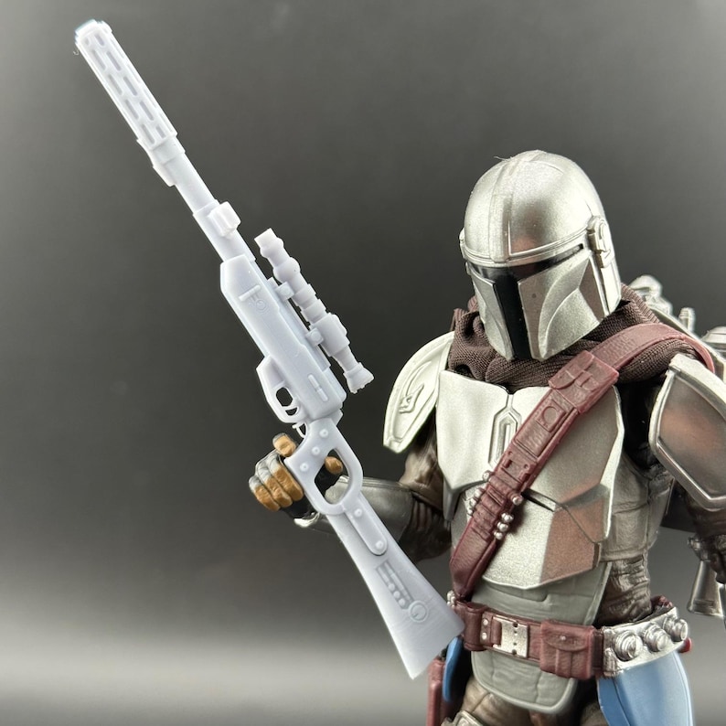 Puede incluir: Una figura de acci&oacute;n mandaloriana gris claro que sostiene un rifle gris claro con mira telesc&oacute;pica. La figura lleva un casco plateado, una bufanda marr&oacute;n y una placa pectoral marr&oacute;n y plateada. La figura tiene un cintur&oacute;n marr&oacute;n y un atuendo azul y marr&oacute;n.