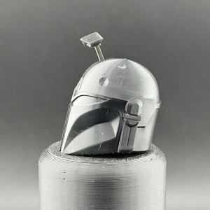 K&ouml;nnte beinhalten: Ein detaillierter, metallisch grauer Mandalorianer-Helm mit einem markanten Visier und einer Antenne. Der Helm ist vor einem neutralen Hintergrund platziert und zeigt sein kompliziertes Design und sein kampferprobtes Aussehen. Es ist ein Sammlerst&uuml;ck.