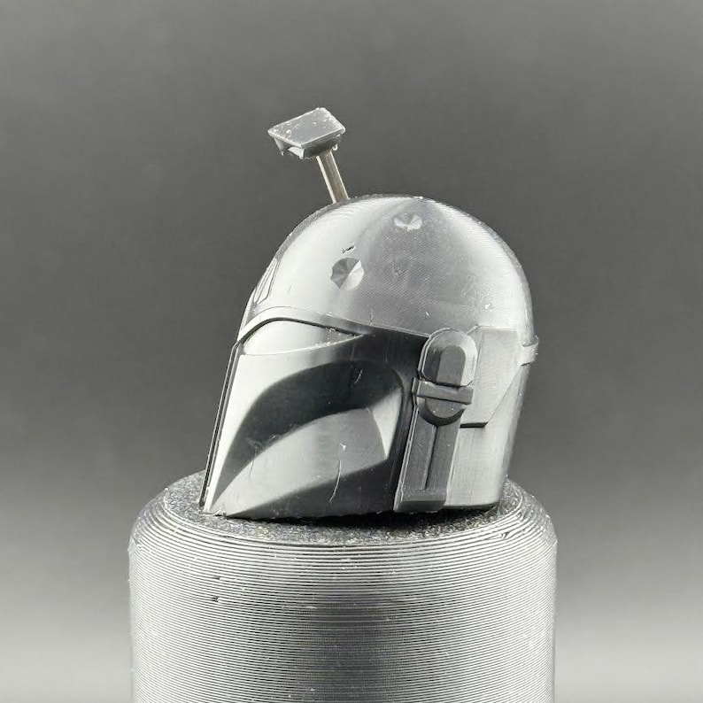 K&ouml;nnte beinhalten: Ein detaillierter, metallisch grauer Mandalorianer-Helm mit einem markanten Visier und einer Antenne. Der Helm ist vor einem neutralen Hintergrund platziert und zeigt sein kompliziertes Design und sein kampferprobtes Aussehen. Es ist ein Sammlerst&uuml;ck.
