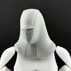 Peut inclure: Une figurine d'action grise claire, repr&eacute;sentant la t&ecirc;te et le haut du torse. La t&ecirc;te est dot&eacute;e d'un design &agrave; capuche avec une visi&egrave;re et une surface textur&eacute;e d&eacute;taill&eacute;e. Les &eacute;paules et la poitrine de la figurine sont blanches, avec des d&eacute;tails subtils.