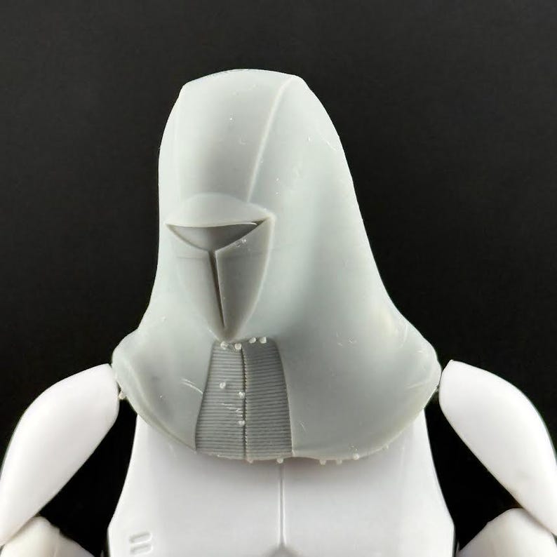 Peut inclure: Une figurine d'action grise claire, repr&eacute;sentant la t&ecirc;te et le haut du torse. La t&ecirc;te est dot&eacute;e d'un design &agrave; capuche avec une visi&egrave;re et une surface textur&eacute;e d&eacute;taill&eacute;e. Les &eacute;paules et la poitrine de la figurine sont blanches, avec des d&eacute;tails subtils.