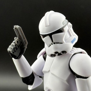May include: A white action figure of a Star Wars clone trooper holding a black blaster pistol. The figure has a white helmet with blue markings and a white body suit.