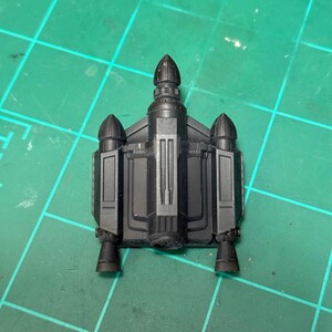 以下が含まれることがあります： 黒色のミニチュアジェットパックモデル。2つのサイドブースターと中央のフィンなど、詳細な特徴があります。ジェットパックはマットな素材でできており、緑色のグリッド面に置かれています。