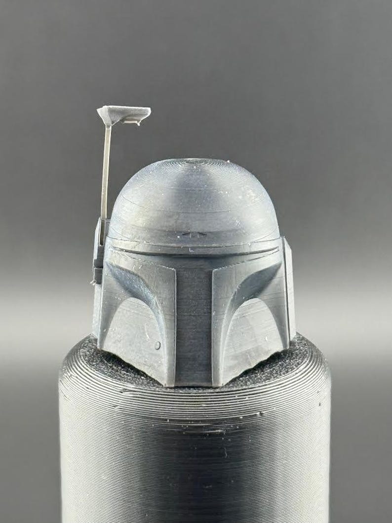 Pu&ograve; includere: Un modello grigio di un casco di Boba Fett stampato in 3D con un'antenna rialzata.