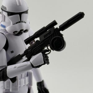 以下が含まれることがあります： 白いストームトルーパーのアクションフィギュアが黒いブラスターライフルを持っています。フィギュアは白いヘルメットとアーマーを着用しています。ブラスターライフルにはスコープと円筒形のバレルが付いています。背景は無地の白です。