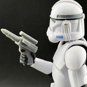 Peut inclure: Figurine d'action blanche repr&eacute;sentant un soldat avec un casque et une armure, tenant un pistolet blaster gris. La figurine a des d&eacute;tails noirs et bleus. Le fond est sombre.