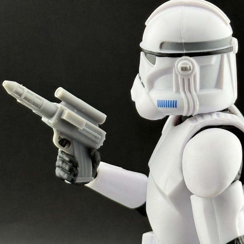 Peut inclure: Figurine d'action blanche repr&eacute;sentant un soldat avec un casque et une armure, tenant un pistolet blaster gris. La figurine a des d&eacute;tails noirs et bleus. Le fond est sombre.