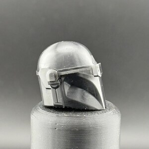 K&ouml;nnte beinhalten: Ein detaillierter, metallisch grauer Mandalorianer-Helm, der das ikonische T-Visier und das markante, kantige Design zeigt. Der Helm befindet sich auf einem strukturierten, zylindrischen Sockel mit dunklem Hintergrund.