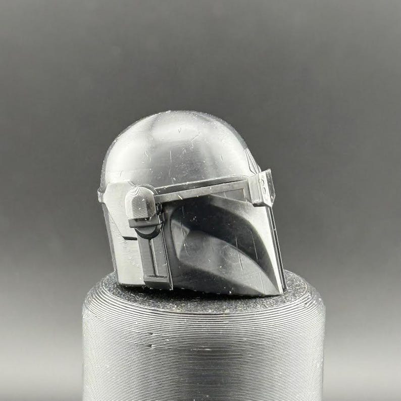 K&ouml;nnte beinhalten: Ein detaillierter, metallisch grauer Mandalorianer-Helm, der das ikonische T-Visier und das markante, kantige Design zeigt. Der Helm befindet sich auf einem strukturierten, zylindrischen Sockel mit dunklem Hintergrund.