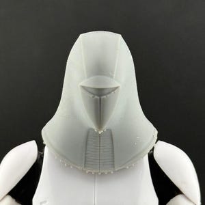 Peut inclure: Gros plan sur un casque en plastique gris avec une visi&egrave;re pointue et une surface textur&eacute;e. Le casque fait partie d'un costume plus large, avec une armure d'&eacute;paule blanche visible en dessous. Le fond est noir uni.