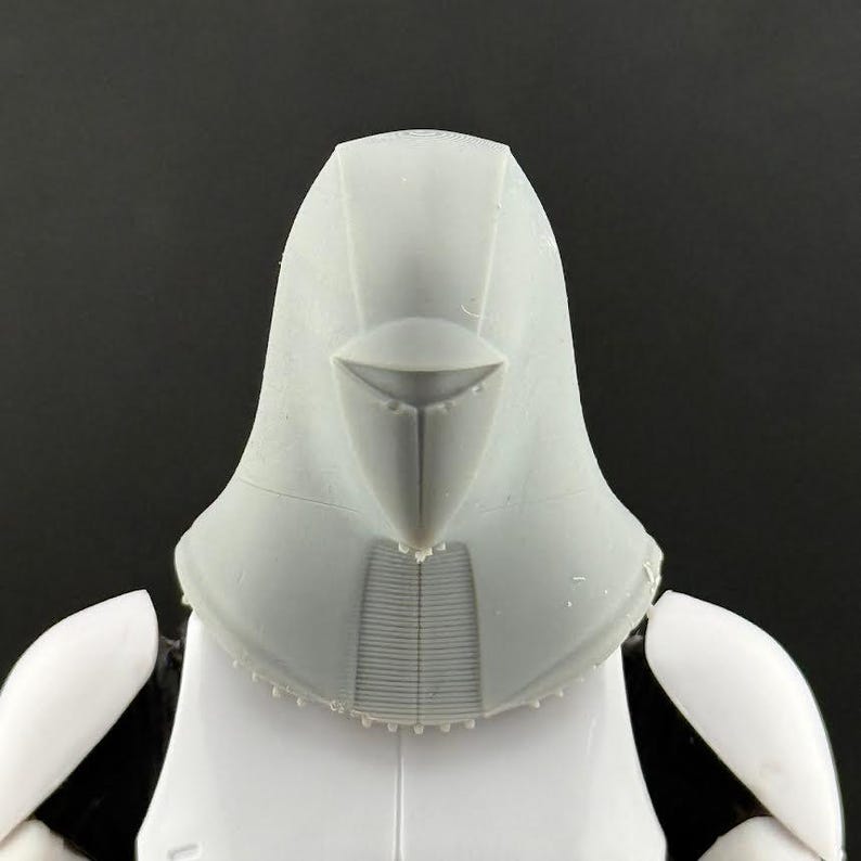 Peut inclure: Gros plan sur un casque en plastique gris avec une visi&egrave;re pointue et une surface textur&eacute;e. Le casque fait partie d'un costume plus large, avec une armure d'&eacute;paule blanche visible en dessous. Le fond est noir uni.