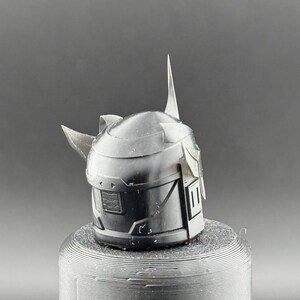 Puede incluir: Un casco gris oscuro detallado con caracter&iacute;sticas angulares, incluyendo una prominente espiga central y protuberancias laterales. El casco est&aacute; montado sobre una base cil&iacute;ndrica texturizada. El dise&ntilde;o evoca un tema futurista o de fantas&iacute;a.