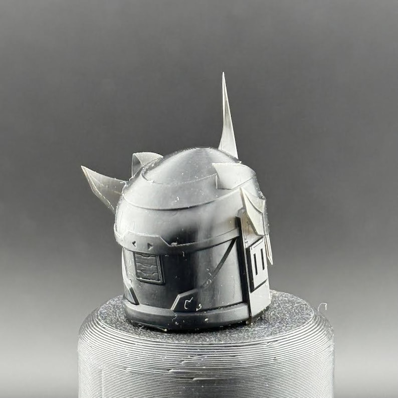 Puede incluir: Un casco gris oscuro detallado con caracter&iacute;sticas angulares, incluyendo una prominente espiga central y protuberancias laterales. El casco est&aacute; montado sobre una base cil&iacute;ndrica texturizada. El dise&ntilde;o evoca un tema futurista o de fantas&iacute;a.