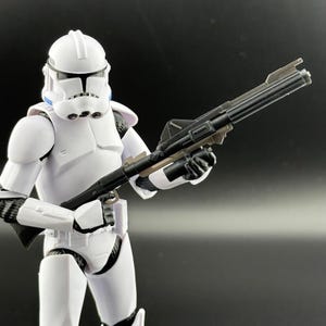 以下が含まれることがあります： 白いスターウォーズのクローン・トルーパーのアクションフィギュアが、黒いブラスターライフルを持っています。トルーパーは、青いマークのついた白いヘルメットをかぶっています。