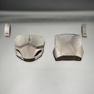 Pu&ograve; includere: Un set di quattro pezzi di armatura grigio scuro, tra cui due corazze e due componenti rettangolari pi&ugrave; piccoli. Le corazze presentano un design angolare dettagliato con cuciture e ritagli visibili. I pezzi sono disposti su una superficie riflettente.