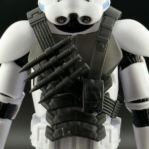 May include: A white and black action figure of a character in a helmet and armour. The figure has a black chest plate with several black cylindrical objects attached. The helmet has black eye lenses and a blue stripe.